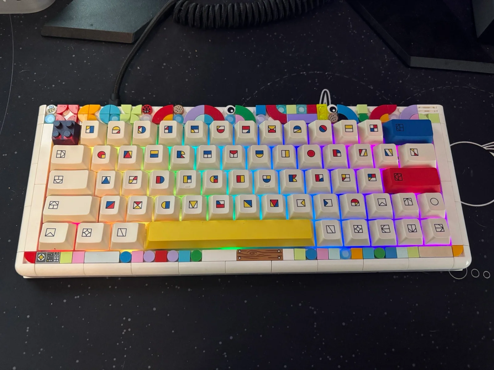 White 65% keyboard with colorful LEGO-themed keycaps featuring brick icons, a bright yellow spacebar, red enter key, and rainbow RGB backlighting on a dark desk