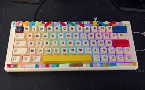 White 65% keyboard with colorful LEGO-themed keycaps featuring brick icons, a bright yellow spacebar, red enter key, and rainbow RGB backlighting on a dark desk