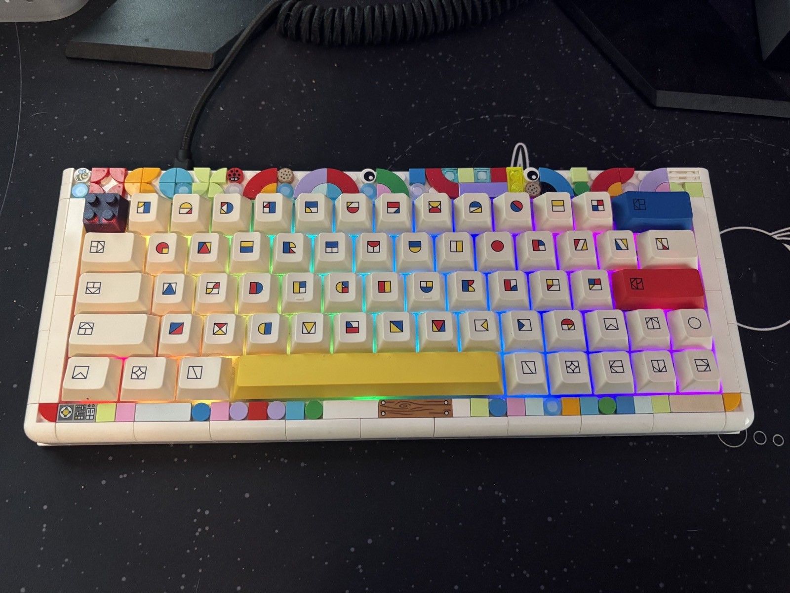 White 65% keyboard with colorful LEGO-themed keycaps featuring brick icons, a bright yellow spacebar, red enter key, and rainbow RGB backlighting on a dark desk