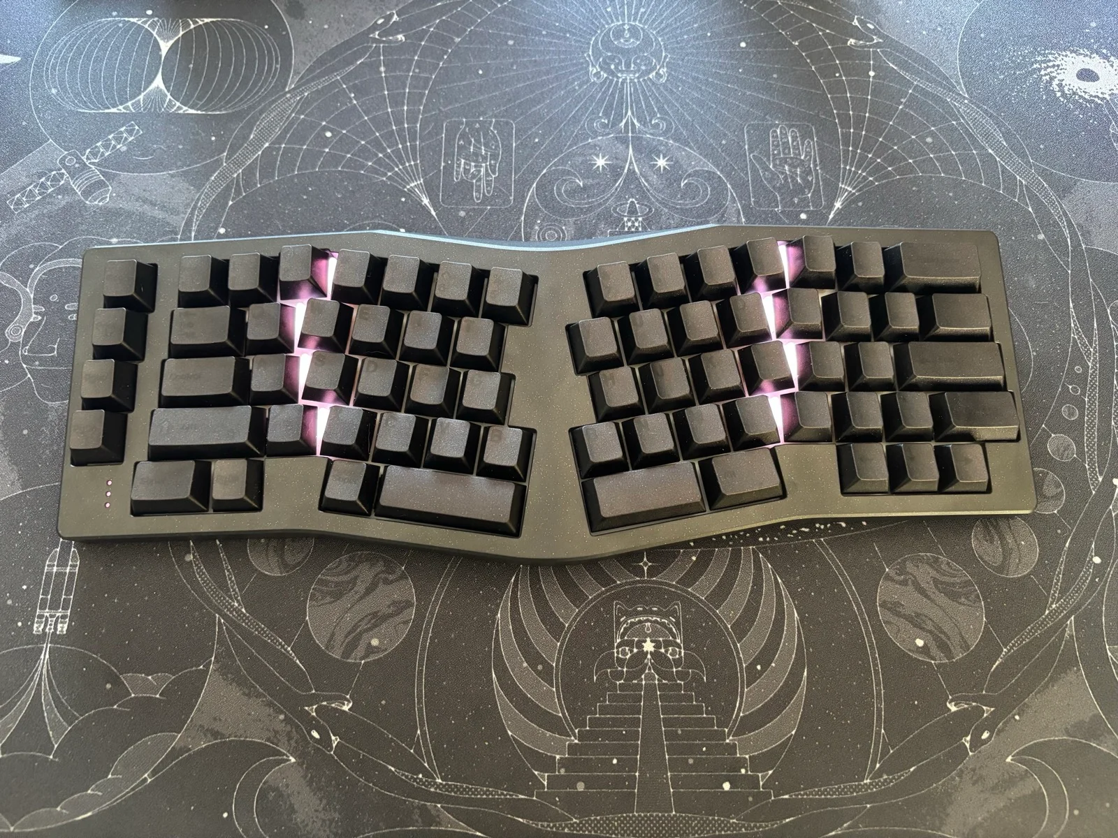 Dark ergonomic split keyboard with black keycaps and purple RGB backlighting on a cosmic-themed deskmat