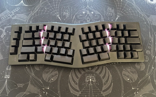 Dark ergonomic split keyboard with black keycaps and purple RGB backlighting on a cosmic-themed deskmat