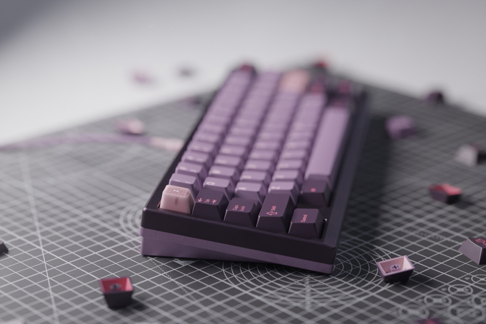 Dark purple 65% keyboard with GMK Symbiote keycaps in deep purple and mauve tones, lighter lavender accent keys, and scattered dark red-purple keycaps on a gray cutting mat