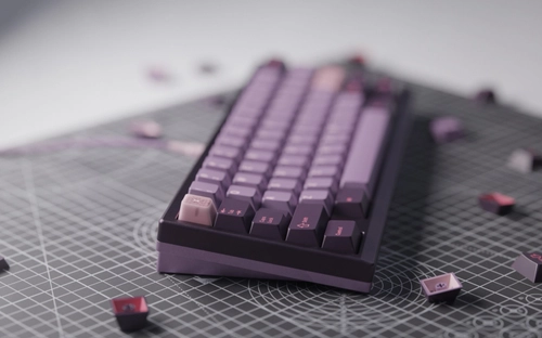 Dark purple 65% keyboard with GMK Symbiote keycaps in deep purple and mauve tones, lighter lavender accent keys, and scattered dark red-purple keycaps on a gray cutting mat